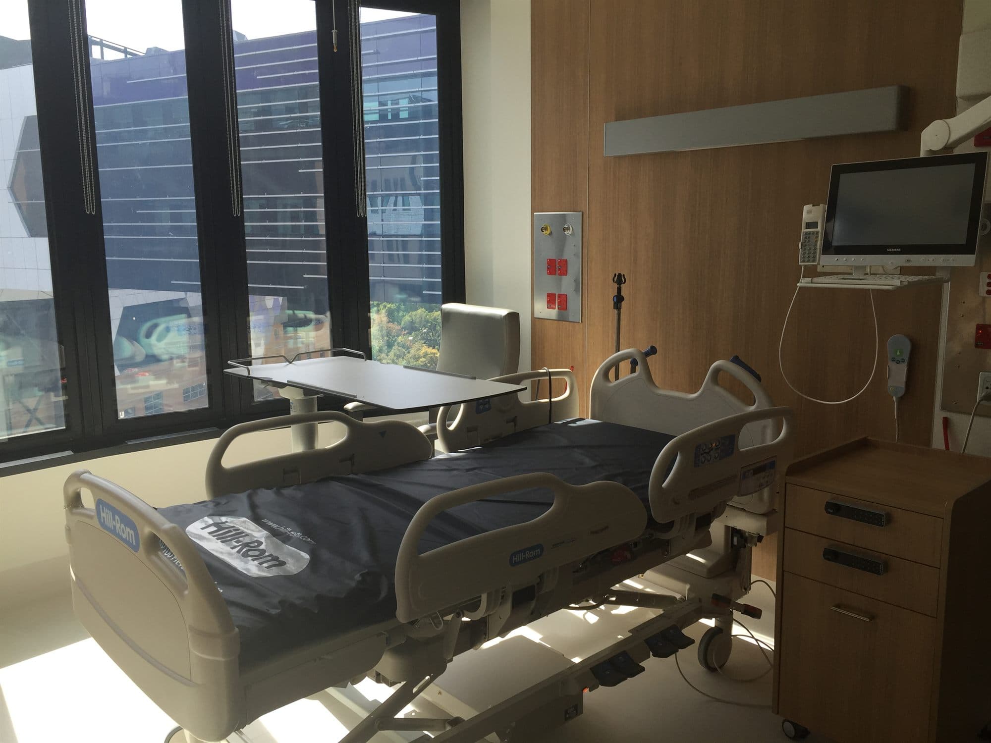 A single patient room with an empty hospital bed, overbed table and bedside monitor beside a large window.