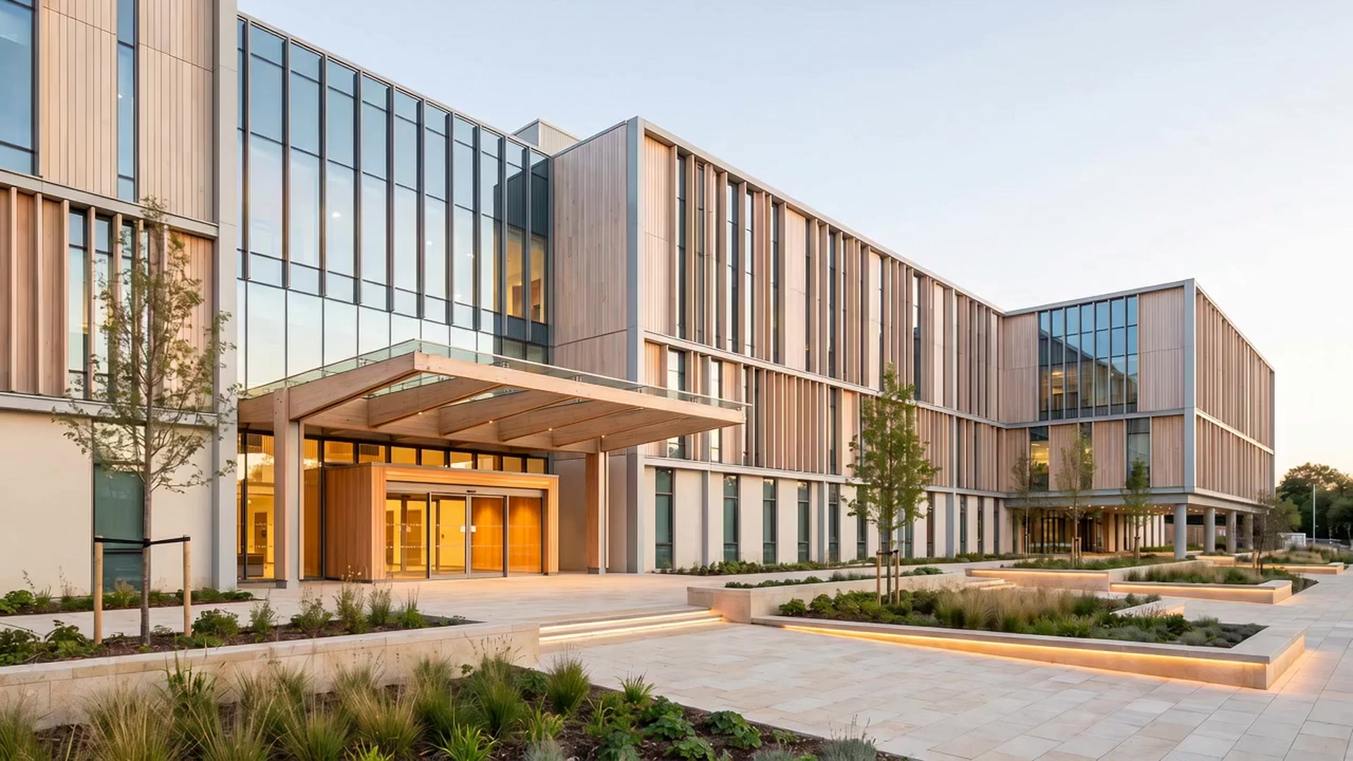 The finished hospital at dusk, with a timber and glass facade and the entrance canopy lit.